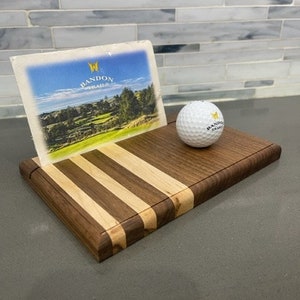 Pu&ograve; includere: Un porta palline da golf in legno con un design a strisce. Il porta palline ha un piccolo slot per le carte e mostra una cartolina con il testo "Bandon Trails". Una pallina da golf bianca con il testo "Bandon Trails" &egrave; appoggiata sul porta palline.