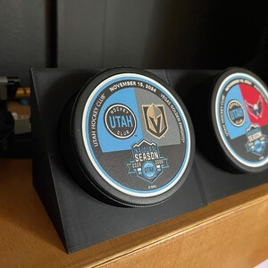May include: Two black hockey pucks with the text "UTAH HOCKEY CLUB" and "VEGAS GOLDEN KNIGHTS" displayed on a black stand. The pucks have blue, gold, and white accents, with the date "NOVEMBER 15, 2024" visible.