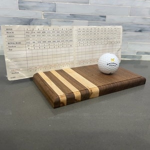 Pu&ograve; includere: Un tagliere di legno con un motivo a strisce di legno chiaro e scuro. Una pallina da golf bianca con il testo "Randox Trials" &egrave; sopra il tagliere. Un pezzo di carta con un tabellone di punteggio del golf &egrave; appoggiato contro il tagliere.