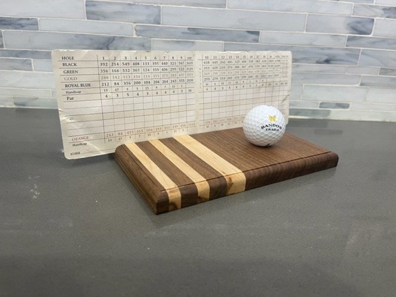 Pu&ograve; includere: Un tagliere di legno con un motivo a strisce di legno chiaro e scuro. Una pallina da golf bianca con il testo "Randox Trials" &egrave; sopra il tagliere. Un pezzo di carta con un tabellone di punteggio del golf &egrave; appoggiato contro il tagliere.