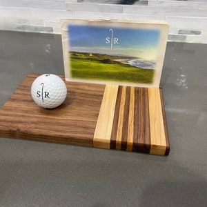Pu&ograve; includere: Un porta palline da golf in legno con un design a strisce. Il porta palline ha una pallina da golf in cima e una cartolina con un'immagine di un campo da golf e il testo "SR" su di essa.