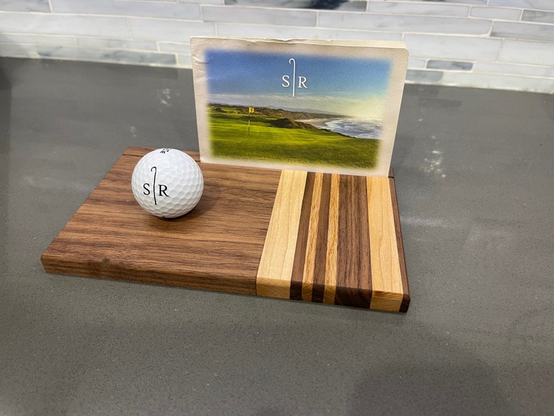 Pu&ograve; includere: Un porta palline da golf in legno con un design a strisce. Il porta palline ha una pallina da golf in cima e una cartolina con un'immagine di un campo da golf e il testo "SR" su di essa.