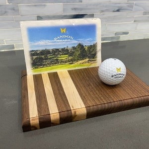 Pu&ograve; includere: Un tagliere di legno con un design a strisce in legno marrone e chiaro. Una palla da golf con le parole "Bandon Trails" stampate su di essa &egrave; appoggiata sul tagliere. Una cartolina con un'immagine di un campo da golf e le parole "Bandon Trails" &egrave; appoggiata contro il tagliere.