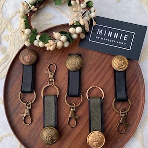 May include: Five keychains with different antique brass buttons and black and green leather straps. The keychains are displayed on a wooden tray. The text "MINNIE BY MONIQUE LUND" is visible on a black card.