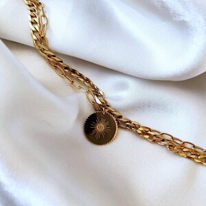 Peut inclure: Un collier en chaîne dorée avec un pendentif rond représentant un soleil avec un œil au centre. La chaîne est une double chaîne avec une chaîne plus épaisse et une chaîne plus fine reliées ensemble.