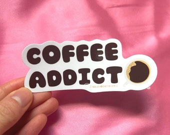 May include: White sticker with black text that reads "COFFEE ADDICT" in a playful font. A small brown coffee cup icon is next to the text. The sticker is held by a hand against a pink background.