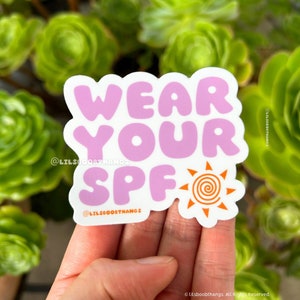 May include: Pink sticker with the text "Wear Your SPF" in a rounded font. A small orange sun with a spiral inside is to the right of the text. The sticker is on a hand.