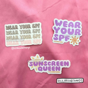 May include: Three stickers with a summer theme. The first sticker is pink and white with the text "Wear Your SPF" repeated four times. The second sticker is purple and white with the text "Wear Your SPF" and a sun graphic. The third sticker is purple and white with the text "Sunscreen Queen" and a sun graphic.
