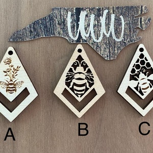 May include: Three wooden diamond-shaped earrings with a bee design. The first earring features a bee with a flower, the second earring features a bee with a honeycomb, and the third earring features a bee with a honeycomb.