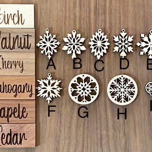 May include: Nine wooden snowflake ornaments in various styles and sizes, labeled A through I. The ornaments are arranged in a row on a wooden surface. The wood types are listed on the left side of the image: Birch, Walnut, Cherry, Mahogany, Sapele, Cedar.