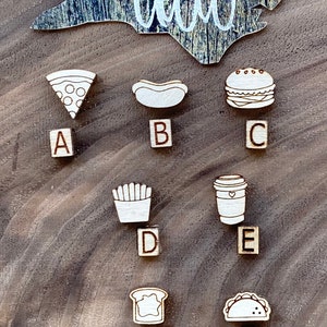 May include: A set of seven wooden food-themed ornaments, each with a letter from A to G. The ornaments are arranged in two rows on a brown wooden surface. The ornaments are painted white and feature various food items, including pizza, hot dog, hamburger, french fries, coffee cup, bread, and taco.