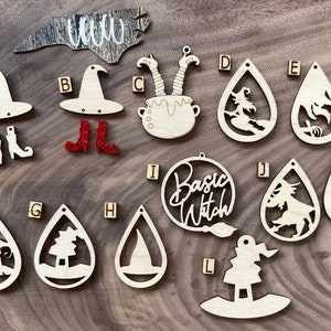 May include: A collection of wooden Halloween-themed cutouts. Includes witch hats, boots, a cauldron, and teardrop-shaped earrings with witch and tree designs. One cutout reads "Basic Witch". The cutouts are made of light-colored wood and red glitter boots.