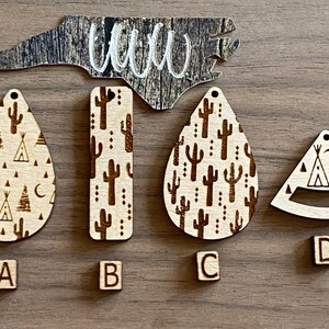 May include: Four wooden laser cut earrings with different designs. The earrings are labeled A, B, C, and D. The designs include a teardrop shape with a cactus and tent pattern, a rectangular shape with a cactus pattern, a teardrop shape with a cactus pattern, and a triangle shape with a tent and moon pattern.
