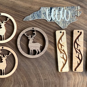 May include: A set of wooden ornaments featuring deer silhouettes and a North Carolina state outline. The ornaments are laser cut and have a natural wood finish. There are also three rectangular wooden ornaments with a branch design.
