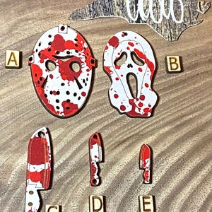 May include: A set of five wooden ornaments featuring horror movie characters and weapons. The ornaments are painted white with red blood splatters. The characters include a hockey mask, a ghost face mask, and a knife. The ornaments are labeled with the letters A, B, C, D, and E.