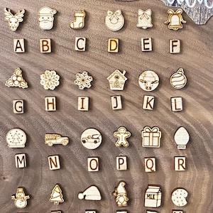 May include: A set of 26 wooden ornaments with Christmas-themed images and letters A through X. The ornaments are arranged in four rows on a wooden surface.
