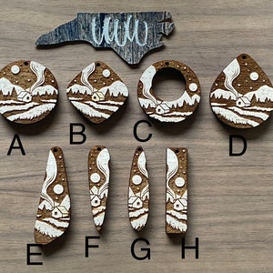 May include: Eight wooden ornaments with a white cabin and snowy landscape design. The ornaments are in various shapes, including circles, ovals, and rectangles. The ornaments are labeled with letters A through H.