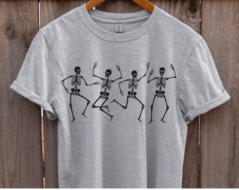 May include: A grey t-shirt featuring a graphic of four skeletons dancing. The skeletons are black and are in a line, facing forward. The shirt has a crew neck and short sleeves.