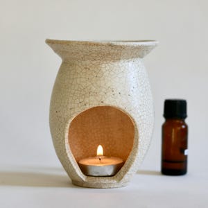 May include: A cream-colored ceramic oil burner with a distinct crackle glaze finish. The burner features a wide top basin and a large arched opening at the front containing a lit tea light candle. A small brown glass essential oil bottle sits nearby.