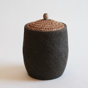 以下が含まれることがあります： 編まれた蓋が付いた黒い陶器の壺。壺はざらざらとした質感の表面をしています。