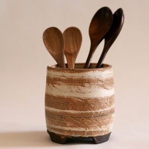 May include: A white ceramic utensil holder with brown streaks and a textured finish. Four wooden spoons are standing upright in the holder.