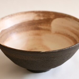 May include: A dark brown ceramic bowl with a white interior. The bowl has a rough, textured exterior and a smooth, white interior. The bowl is slightly wider at the top than at the bottom.