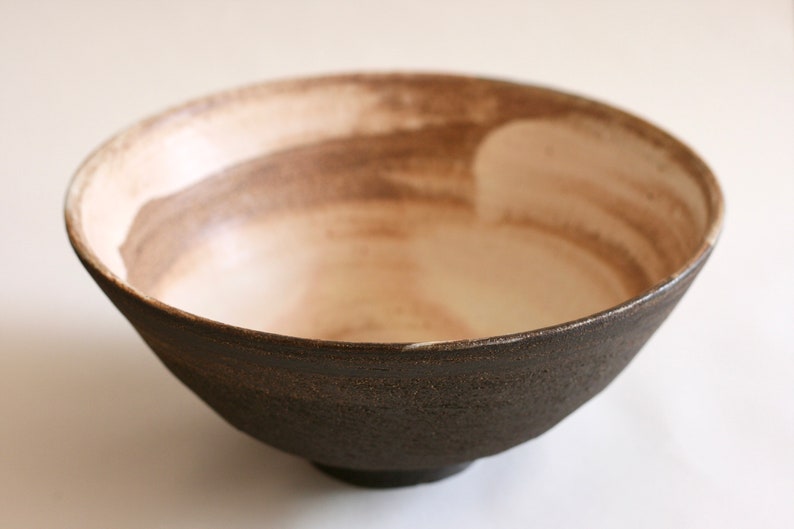 May include: A dark brown ceramic bowl with a white interior. The bowl has a rough, textured exterior and a smooth, white interior. The bowl is slightly wider at the top than at the bottom.