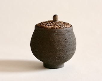 Op de afbeelding: Een donkerbruine keramische pot met een deksel van geweven bruin materiaal. Het deksel heeft een kleine knop bovenop.