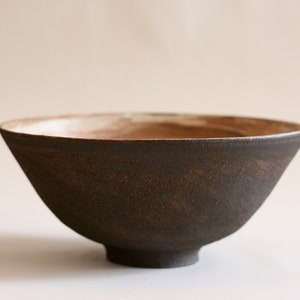 May include: A dark brown ceramic bowl with a slightly raised base. The bowl has a rough, textured surface and a slightly lighter brown rim.