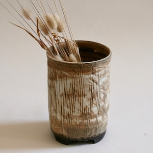 May include: A white ceramic vase with a brown glaze and a textured surface. The vase has a cylindrical shape and a wide opening. It is sitting on a white surface and has dried grasses inside.