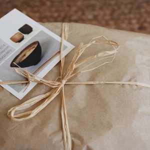 May include: A brown paper package tied with a natural twine bow. A business card with the text "Tom Tzu Pottery" and contact information is on top of the package.
