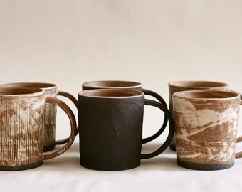 May include: Six ceramic mugs with different brown and white glaze patterns. The mugs have rounded handles and are arranged in a row on a white surface.