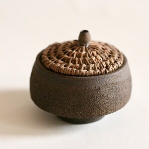 Peut inclure: Un petit pot en céramique brun foncé avec un couvercle en osier. Le pot a une surface rugueuse et texturée.