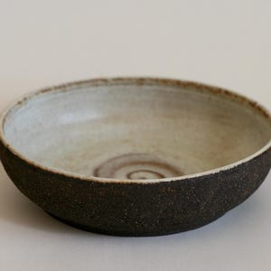 Op de afbeelding: Een witte keramische kom met een donkerbruine rand en een subtiel swirl-patroon aan de binnenkant. De kom heeft een ruwe, getextureerde buitenkant.