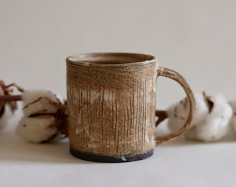 Puede incluir: Una taza de cerámica hecha a mano con un diseño texturizado de líneas verticales en tonos marrones y beige. La taza tiene una base marrón oscuro y un asa curva. Ramas de algodón en el fondo.