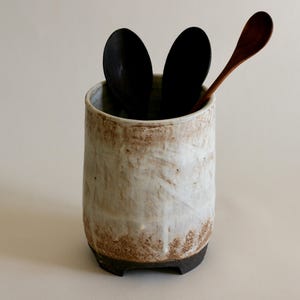 Op de afbeelding: Een witte keramische bestekhouder met een bruine glazuur en zwarte voetjes. De houder bevat twee zwarte lepels en een houten lepel.