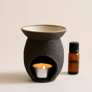 Pode incluir: Um difusor de óleo de cerâmica preta com uma borda branca e uma pequena vela de chá dentro. Uma pequena garrafa de vidro marrom de óleo essencial está ao lado do difusor.