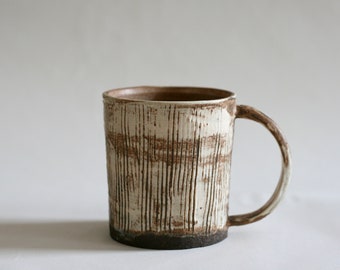 May include: A handmade ceramic mug with a textured, vertical-lined design. The mug features a white and brown color scheme, with a dark brown base and handle. The mug is approximately 4 inches tall.