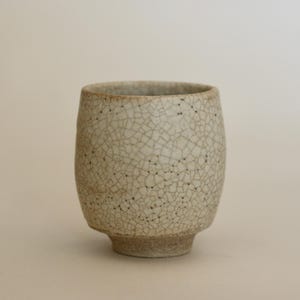 Puede incluir: Una taza de cerámica hecha a mano con un esmalte craquelado único. La taza es de color beige claro con una red de finas grietas y pequeños puntos oscuros. Tiene un cuerpo redondeado y una base pequeña, ligeramente ensanchada.