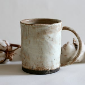 Puede incluir: Una taza de cerámica blanca con un borde y un asa marrones. La taza tiene un aspecto rústico y artesanal y es perfecta para disfrutar de una bebida caliente.