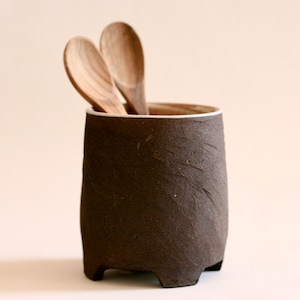 Può includere: Un portautensili in ceramica marrone con un bordo bianco e quattro piccoli piedini. Il portautensili è pieno di due cucchiai di legno.