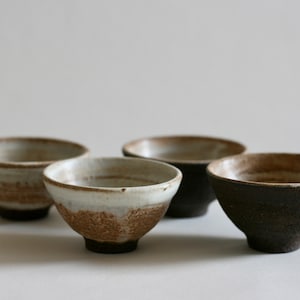 Puede incluir: Cuatro cuencos pequeños de cerámica, dos con un esmalte blanco y detalles marrones, y dos con un esmalte marrón oscuro y detalles marrones.