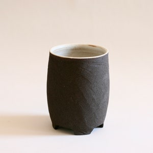 May include: A dark brown ceramic mug with a white interior and a rounded base. The mug has a textured exterior and four small feet.