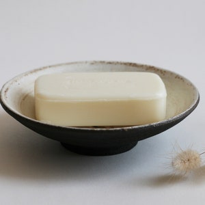 Puede incluir: Una pastilla de jabón blanca se encuentra en un jabonera de cerámica negra y blanca. La jabonera tiene una base elevada y una forma ligeramente ovalada.
