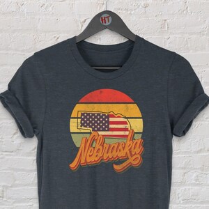 Op de afbeelding: Donkergrijs heather t-shirt met een vintage-stijl grafische afbeelding van de staat Nebraska met een Amerikaanse vlag erin. De afbeelding is geplaatst tegen een zonsondergang achtergrond met het woord "Nebraska" in een retro-lettertype.