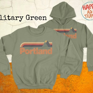 May include: Two sweatshirts in military green, one a crew neck and the other a hoodie, both featuring a retro-style graphic of mountains and the word "Portland". The hoodie has the text "Happy as I Travel" on the back.