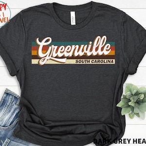 以下が含まれることがあります： ダークグレーヘザーのTシャツに、ヴィンテージスタイルのグラフィックがプリントされています。グラフィックには「グリーンビル、サウスカロライナ」という文字がオレンジ、イエロー、ブルーで書かれています。
