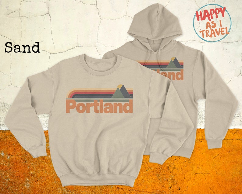 May include: Two sweatshirts in sand colour. One is a crew neck with the text "Portland" and a retro mountain range graphic. The other is a hoodie with the text "land" and a retro mountain range graphic. Both have the text "Happy as I Travel" in a circle.