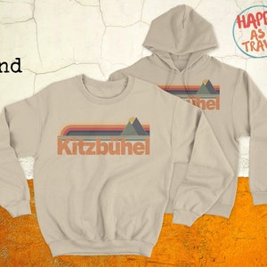 May include: Two sand-coloured sweatshirts with a retro mountain graphic and the text "Kitzbühel". The sweatshirt on the left is a crew neck and the sweatshirt on the right is a hoodie with the text "Happy as I Travel" on the back.
