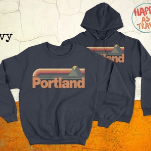 May include: Two navy blue sweatshirts featuring a retro mountain and sunset graphic with the text "Portland". One sweatshirt is a crew neck and the other is a hoodie. The hoodie has the text "Happy as I Travel" on the back.